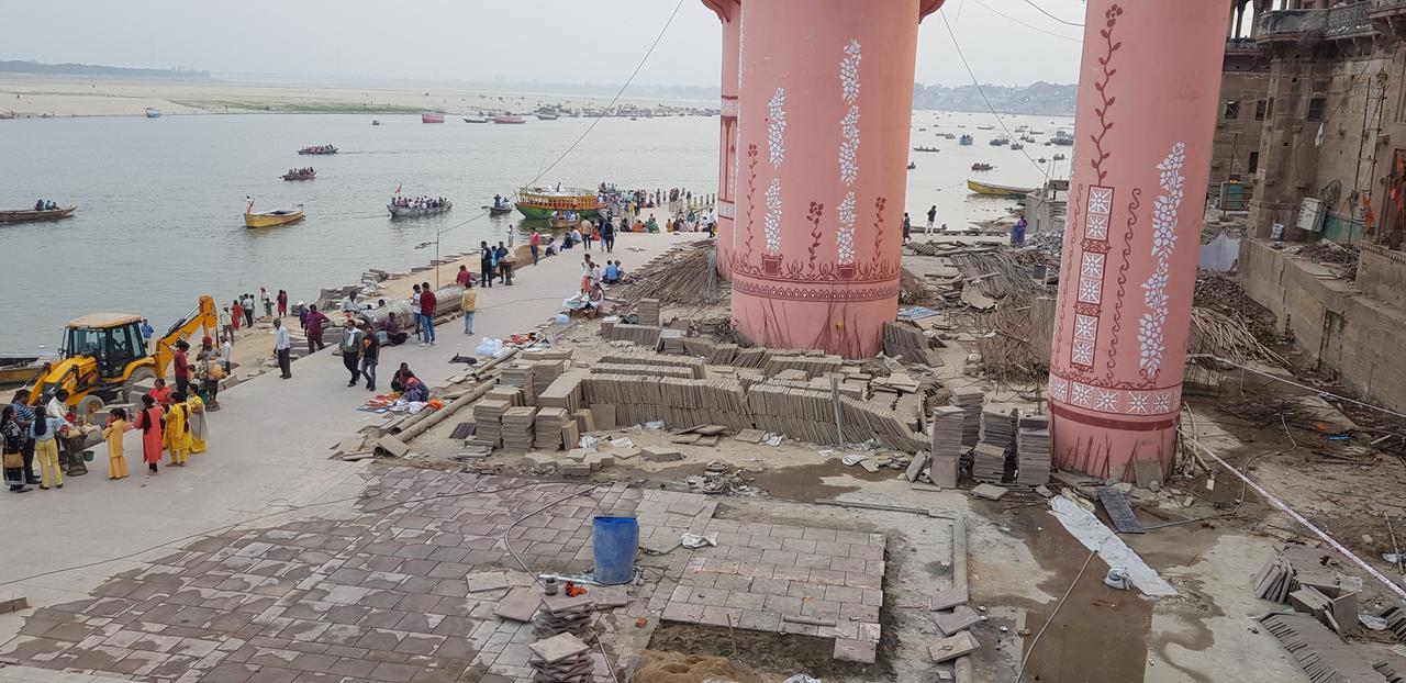 Eine Übersichtsaufnahme von den Bauarbeiten am Lalita Ghat. Auf der linken Seite ist der Ganges zu sehen, auf der rechten Seite erheben sich auf der Baustelle drei große, rosafarbene Säulen empor.