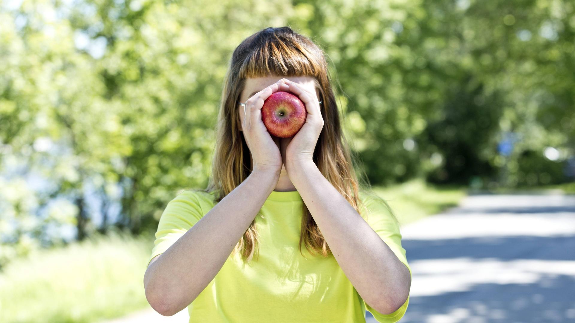 Eine junge Frau hält sich einen Apfel vor ihr Gesicht. (Symbolfoto)