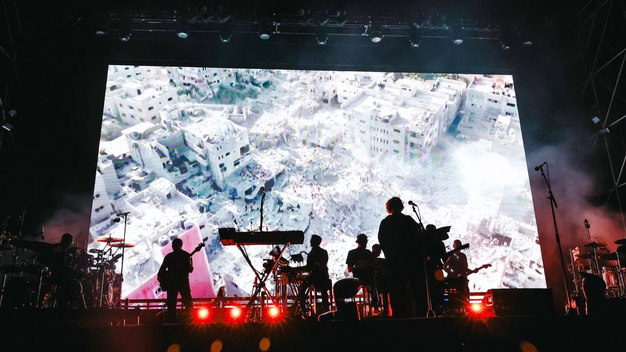 Die Musiker der Band Massive Attack auf der Bühne während eines Auftritts in Mailand, dahinter sind auf einer großen Leinwand völlig zerstörte Gebäude zu sehen