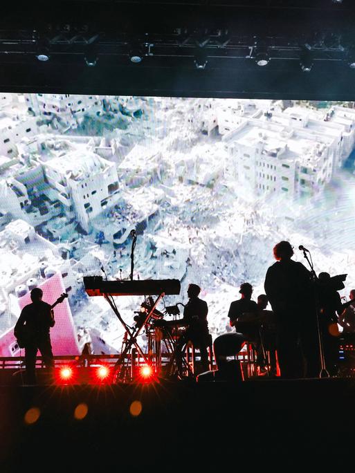 Die Musiker der Band Massive Attack auf der Bühne während eines Auftritts in Mailand, dahinter sind auf einer großen Leinwand völlig zerstörte Gebäude zu sehen