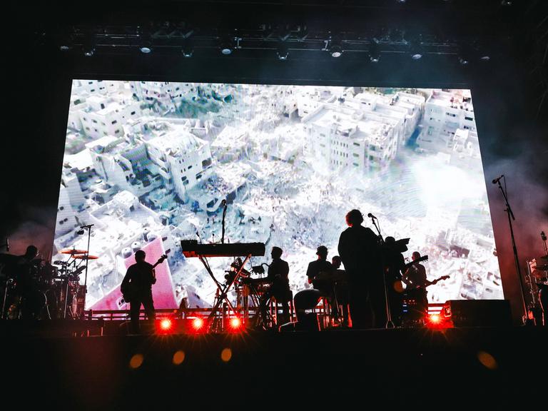 Die Musiker der Band Massive Attack auf der Bühne während eines Auftritts in Mailand, dahinter sind auf einer großen Leinwand völlig zerstörte Gebäude zu sehen