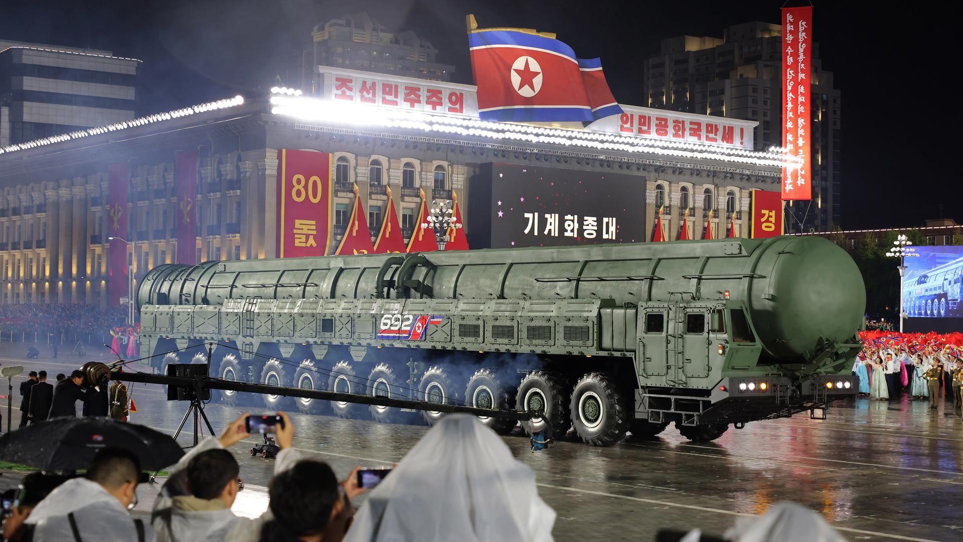 Die Rakete wird auf einem Transporter an einem zum 80. Jahrestag der Arbeiterpartei Nordkoreas rot-gelb geschmückten Haus vorbeigefahren. Es regnet und ist dunkel. Im Vordergrund halten Fotografen in weißen Regencapes Kameras hoch. Die Rakete wird auf einem Transporter an einem zum 80. Jahrestag der Arbeiterpartei Nordkoreas rot-gelb geschmückten Haus vorbeigefahren. Es regnet und ist dunkel. Im Vordergrund halten Fotografen in weißen Regencapes Kameras hoch.