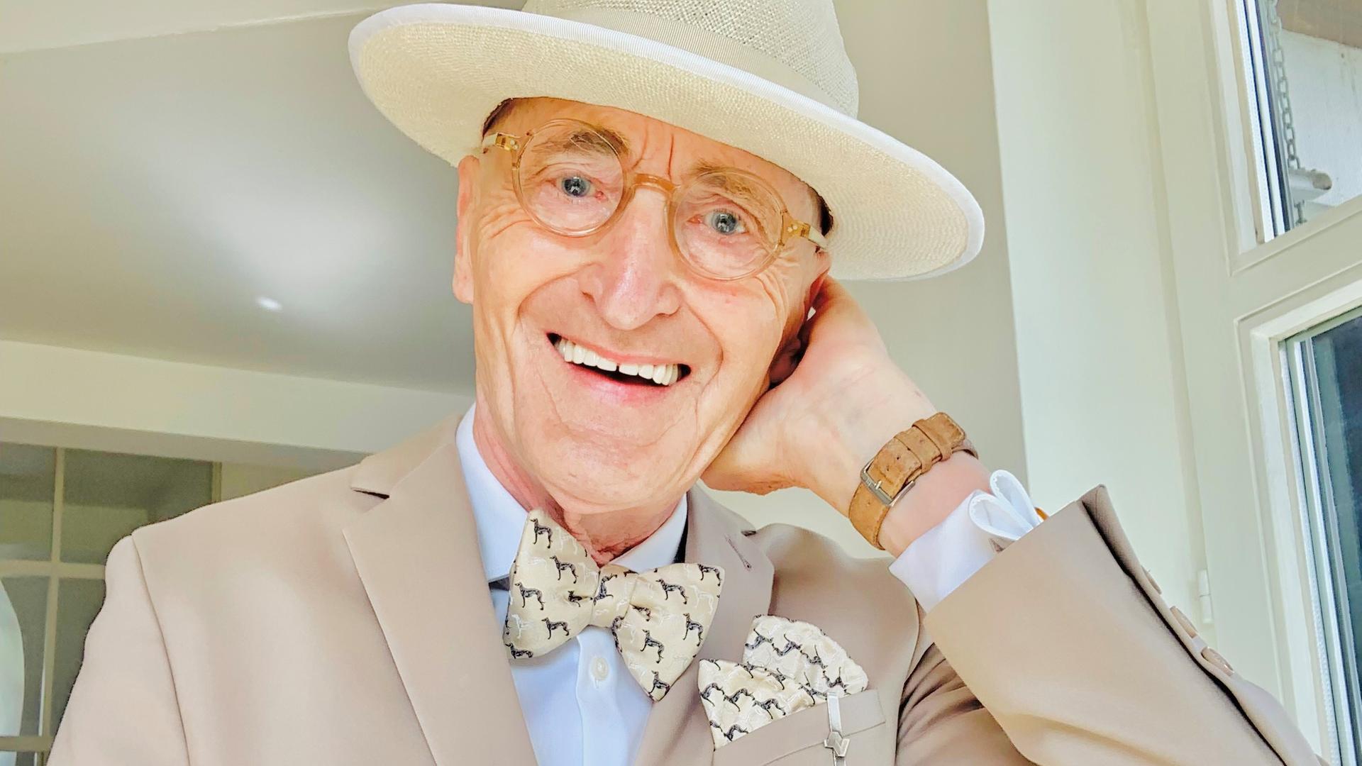 Das Herrenmodel Günther Krabbenhöft, 77, trägt einen hellen Anzug mit Fliege und Hut und lacht in die Kamera.