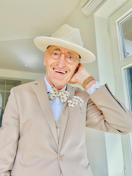 Das Herrenmodel Günther Krabbenhöft, 77, trägt einen hellen Anzug mit Fliege und Hut und lacht in die Kamera.