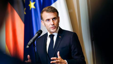 Emmanuel Macron steht vor einem Mikrofon und spricht mir gestikulierenden Händen. Hinter ihm sind die deutsche und die europäische Flagge zu erkennen.
