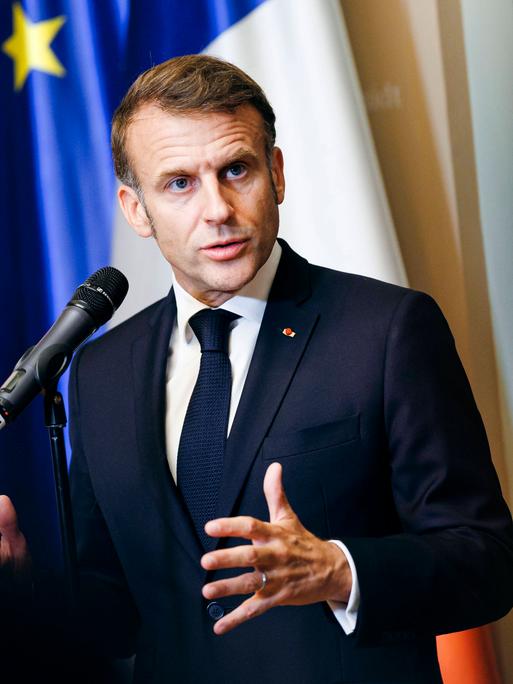 Emmanuel Macron steht vor einem Mikrofon und spricht mir gestikulierenden Händen. Hinter ihm sind die deutsche und die europäische Flagge zu erkennen.