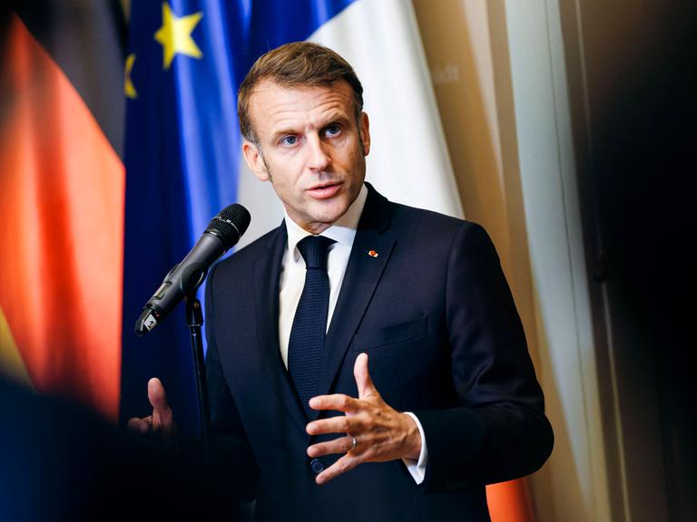 Emmanuel Macron steht vor einem Mikrofon und spricht mir gestikulierenden Händen. Hinter ihm sind die deutsche und die europäische Flagge zu erkennen.