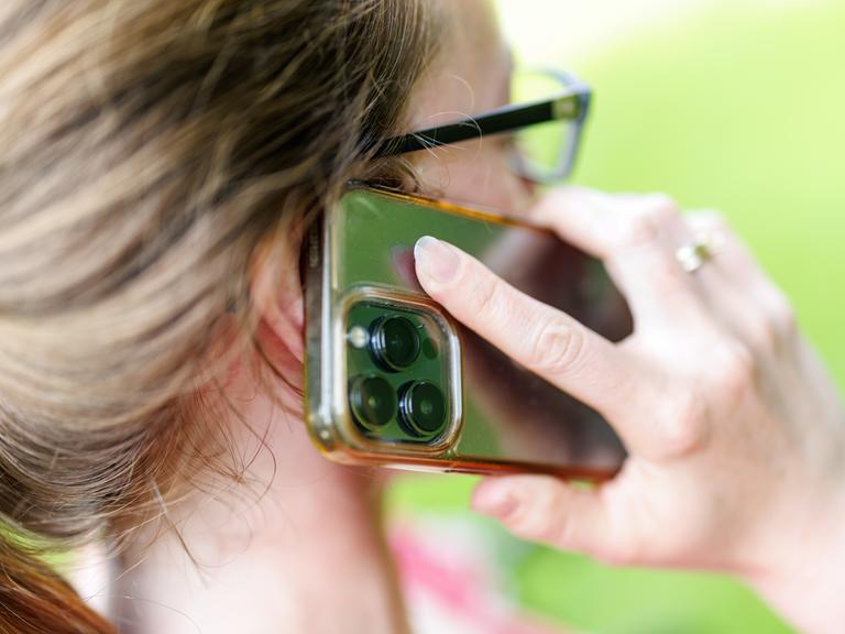 Eine junge Frau steht in einem Garten und telefoniert mit ihrem Smartphone.