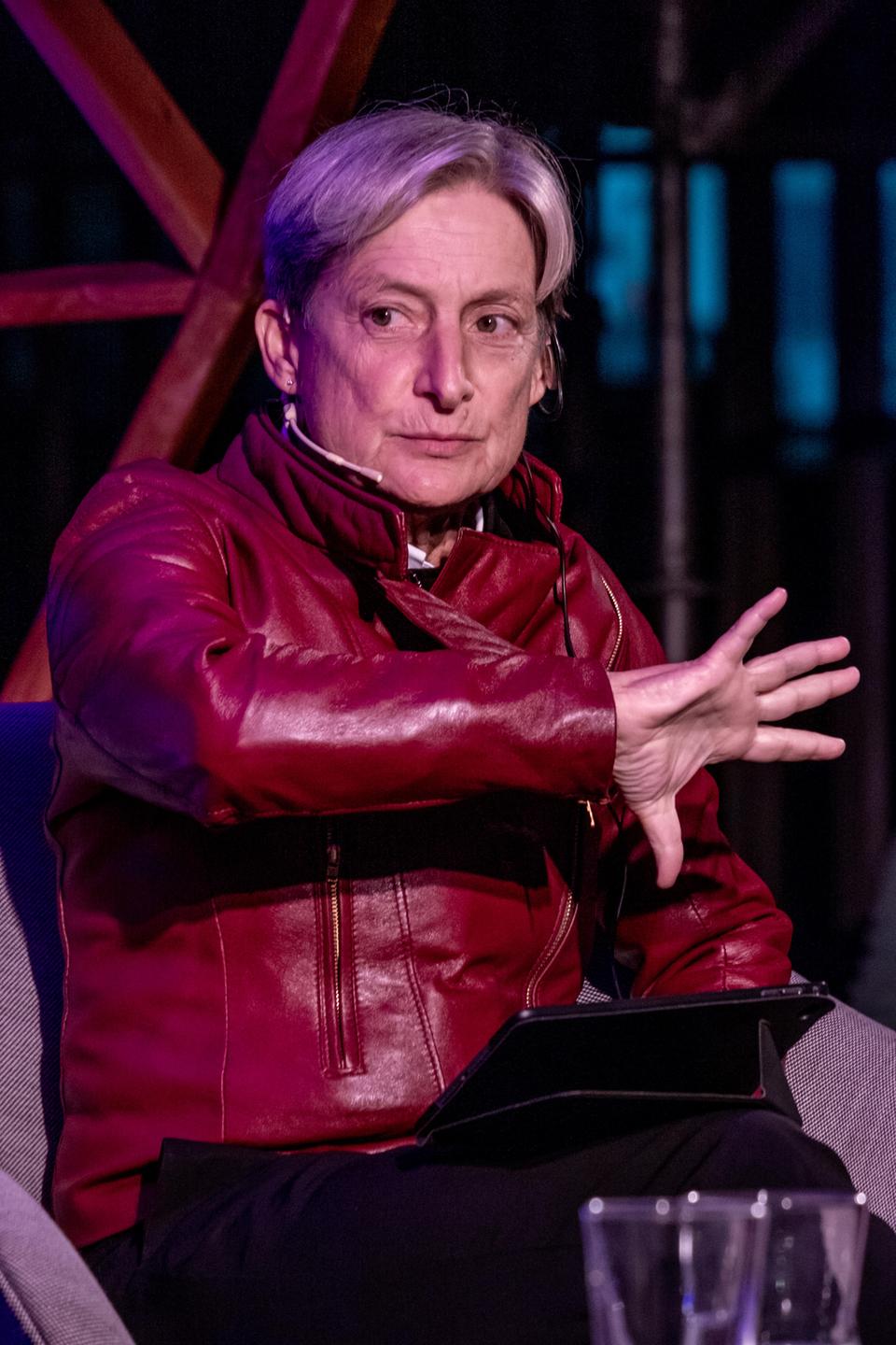 Porträtaufnahme der Philosophin Judith Butler in roter Lederjacke während einer Diskussionsveranstaltung auf einer Bühne.