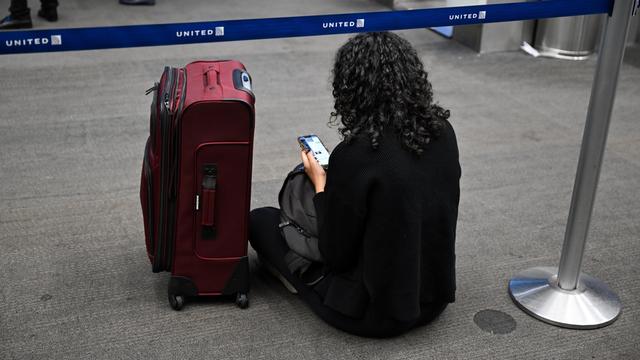 Eine Person sitzt auf dem Boden mit einem Handy in der Hand, daneben steht in roter Koffer.