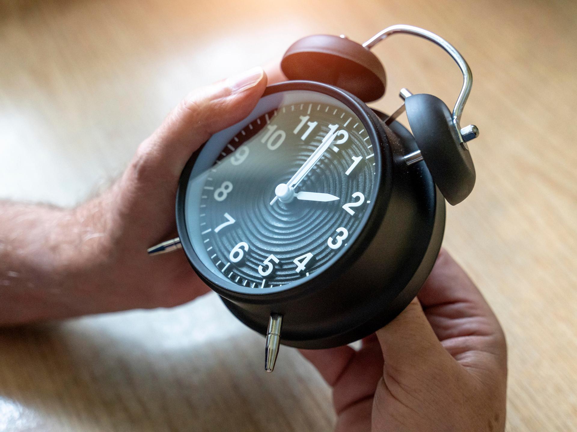 Zwei Hände drehen an einem Wecker, der auf 2 Uhr steht