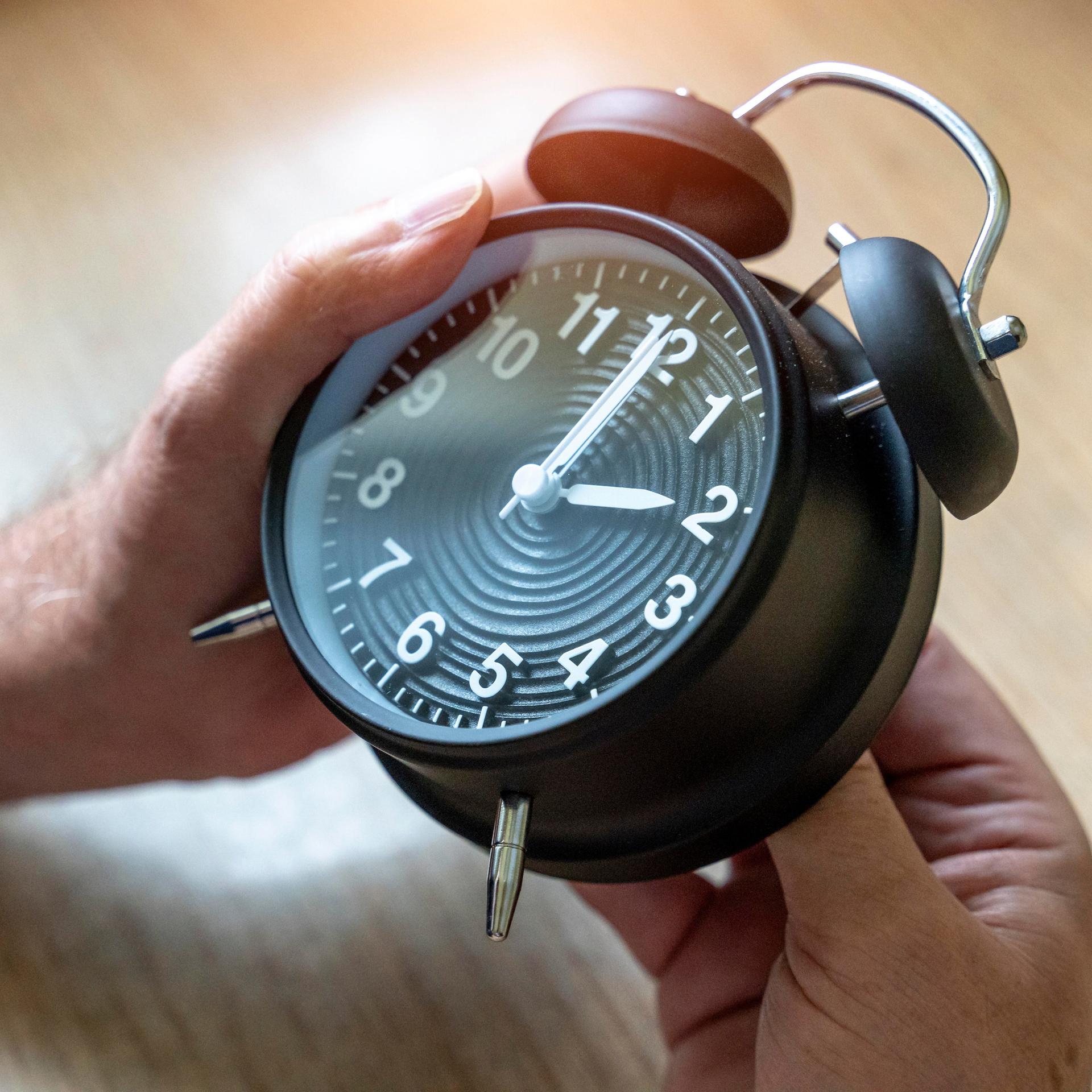 Zwei Hände drehen an einem Wecker, der auf 2 Uhr steht