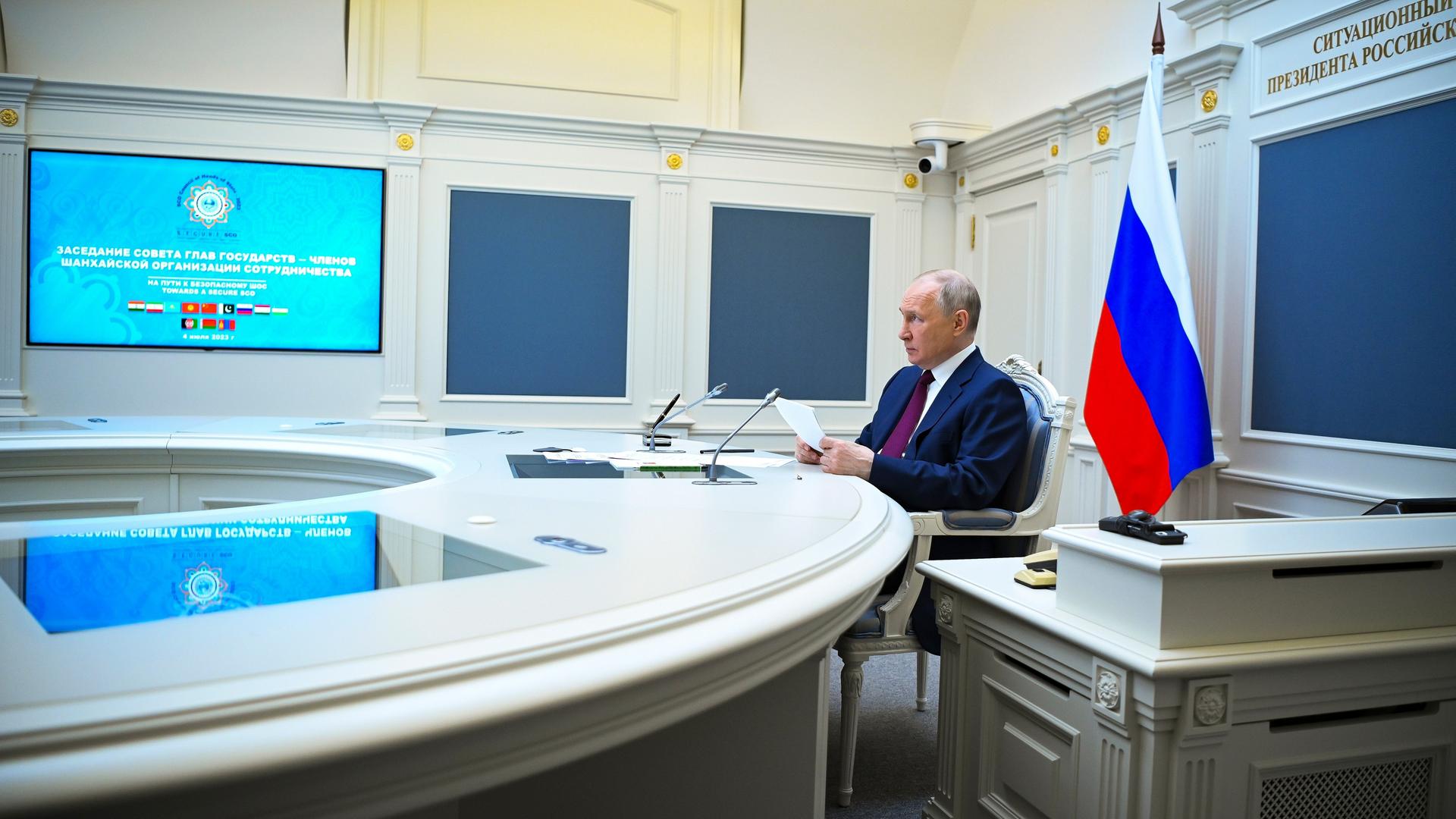 Putin sitzt in einem Raum an einem runden Tisch und hält ein Blatt in beiden Händen. Auf einem Videomonitor sieht man das Logo der Organisation und die Wappen der Mitgliedsstaaten. Neben Putin steht auf einem Schreibtisch eine russische Fahne.