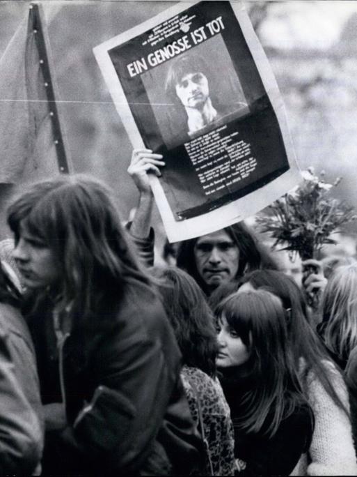 Trauerzug mit Menschen aus dem alternativen Milieu in typischer Kleidung und Anmutung. Einer von ihnen hält ein Schild mit dem Bild von Holger Meins hoch, auf dem steht "Ein Genosse ist tot".