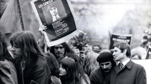 Trauerzug mit Menschen aus dem alternativen Milieu in typischer Kleidung und Anmutung. Einer von ihnen hält ein Schild mit dem Bild von Holger Meins hoch, auf dem steht "Ein Genosse ist tot".