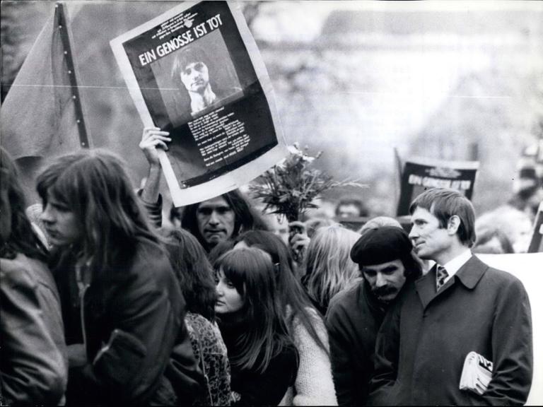 Trauerzug mit Menschen aus dem alternativen Milieu in typischer Kleidung und Anmutung. Einer von ihnen hält ein Schild mit dem Bild von Holger Meins hoch, auf dem steht "Ein Genosse ist tot".