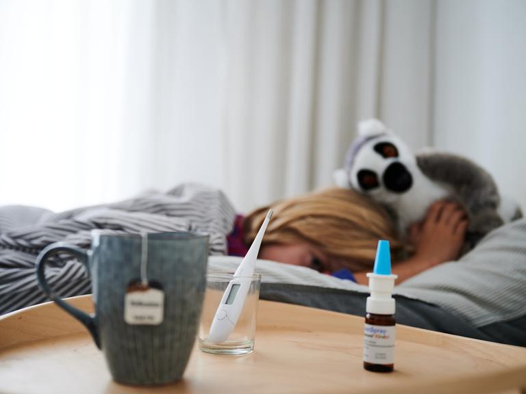 Fieberthermometer, Nasenspray und eine Tasse Tee stehen vor einem Bett, in dem ein Kind mit Kuscheltier liegt.