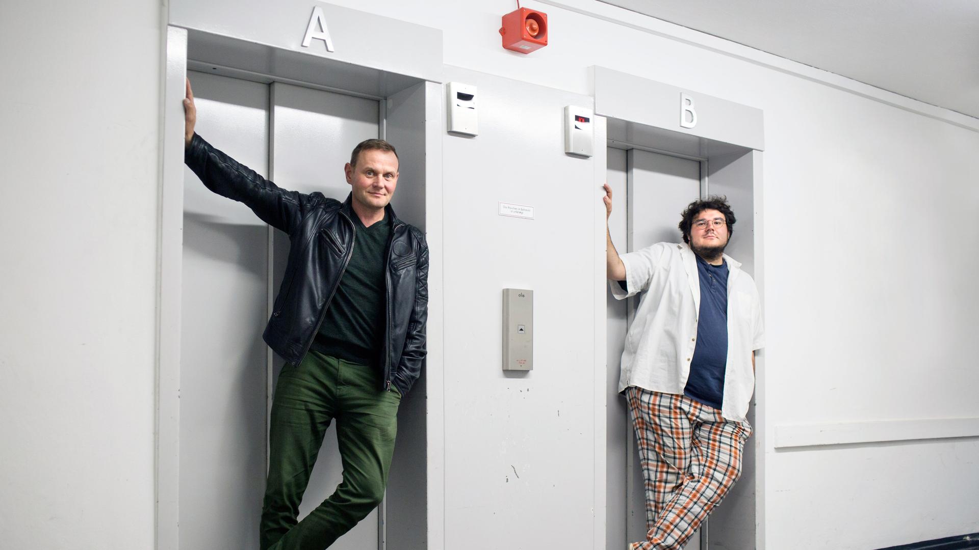 Devid Striesow und Axel Rahnisch stehen jeweils vor einer Fahrstuhltür.