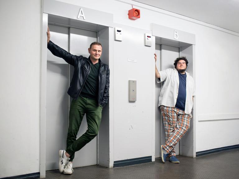Devid Striesow und Axel Rahnisch stehen jeweils vor einer Fahrstuhltür.
