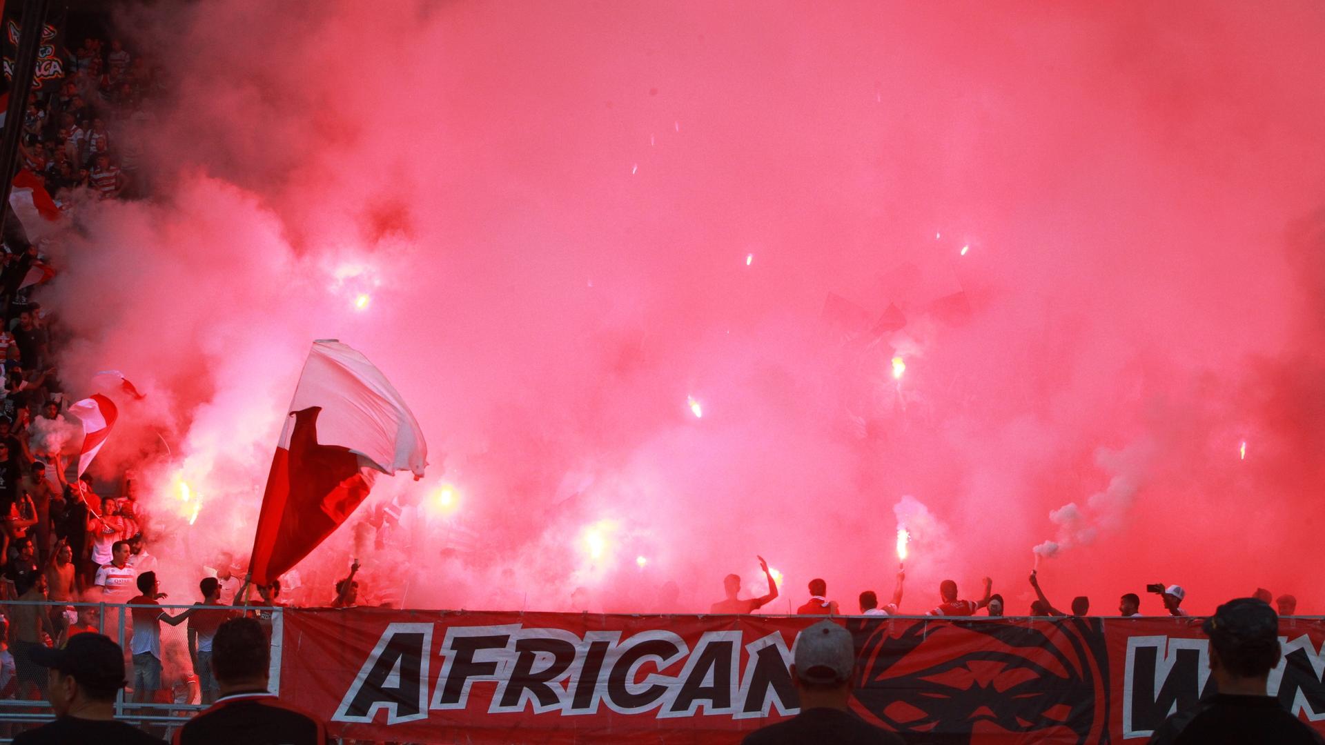 Blick in eine Gruppe Ultras im Stadion, sie sind kaum zu sehen, um sie ist roter Rauch. Am Tribünenrand hängt ein Banner, auf dem African steht.