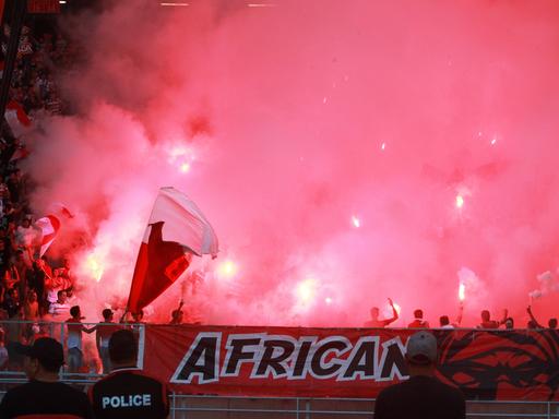 Blick in eine Gruppe Ultras im Stadion, sie sind kaum zu sehen, um sie ist roter Rauch. Am Tribünenrand hängt ein Banner, auf dem African steht.