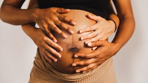 Hände umfassen den Bauch einer schwangeren Person von hinten.