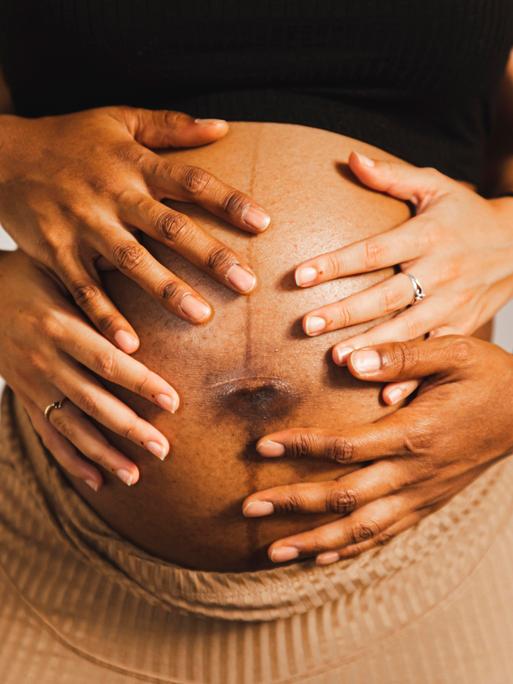 Hände umfassen den Bauch einer schwangeren Person von hinten.