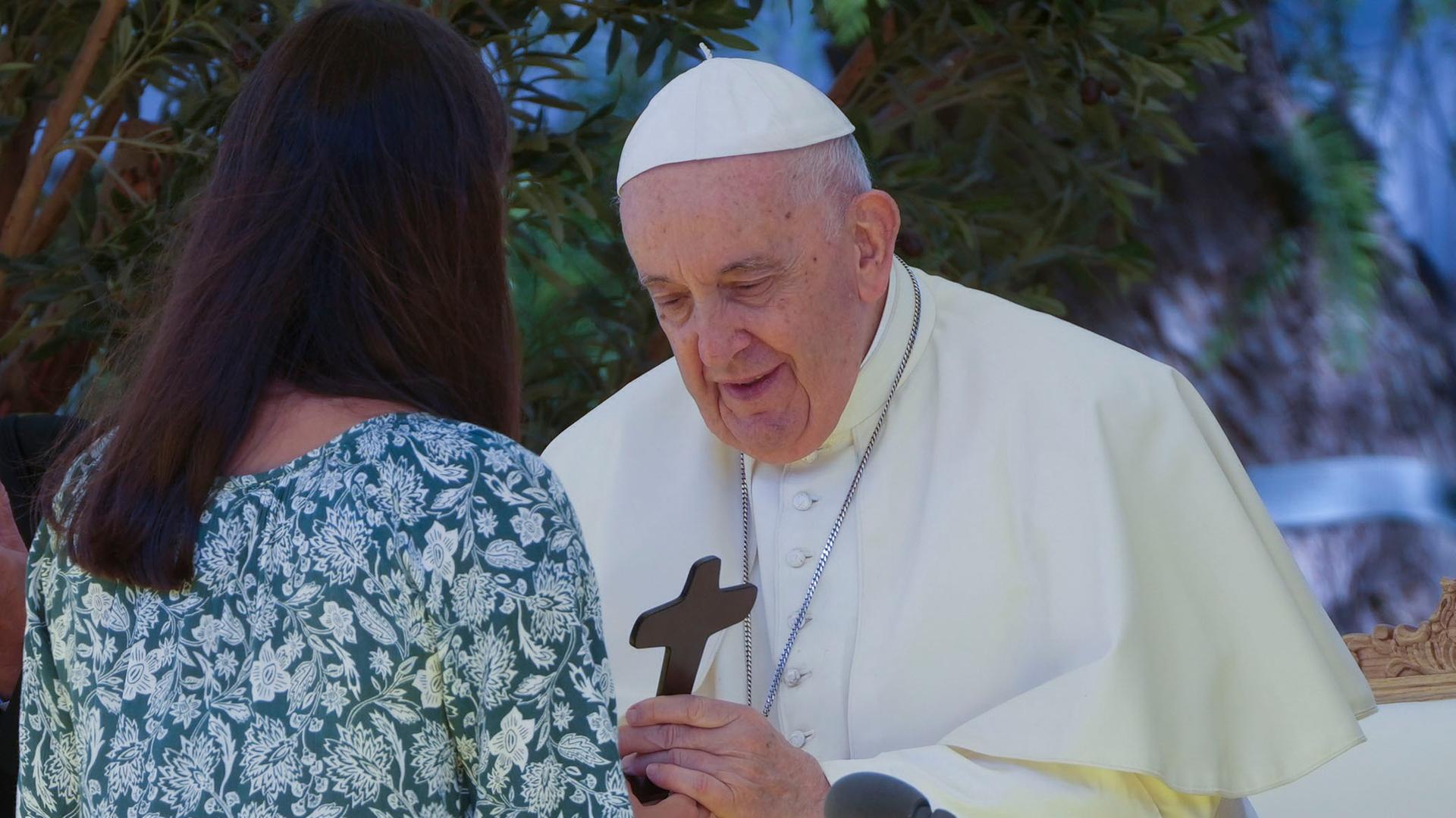 Der Papst im weißen Ornat nimmt von einer Frau im blaugeblümelten Kleid, die man von hinten sieht, ein hölzernes Kreuz an.