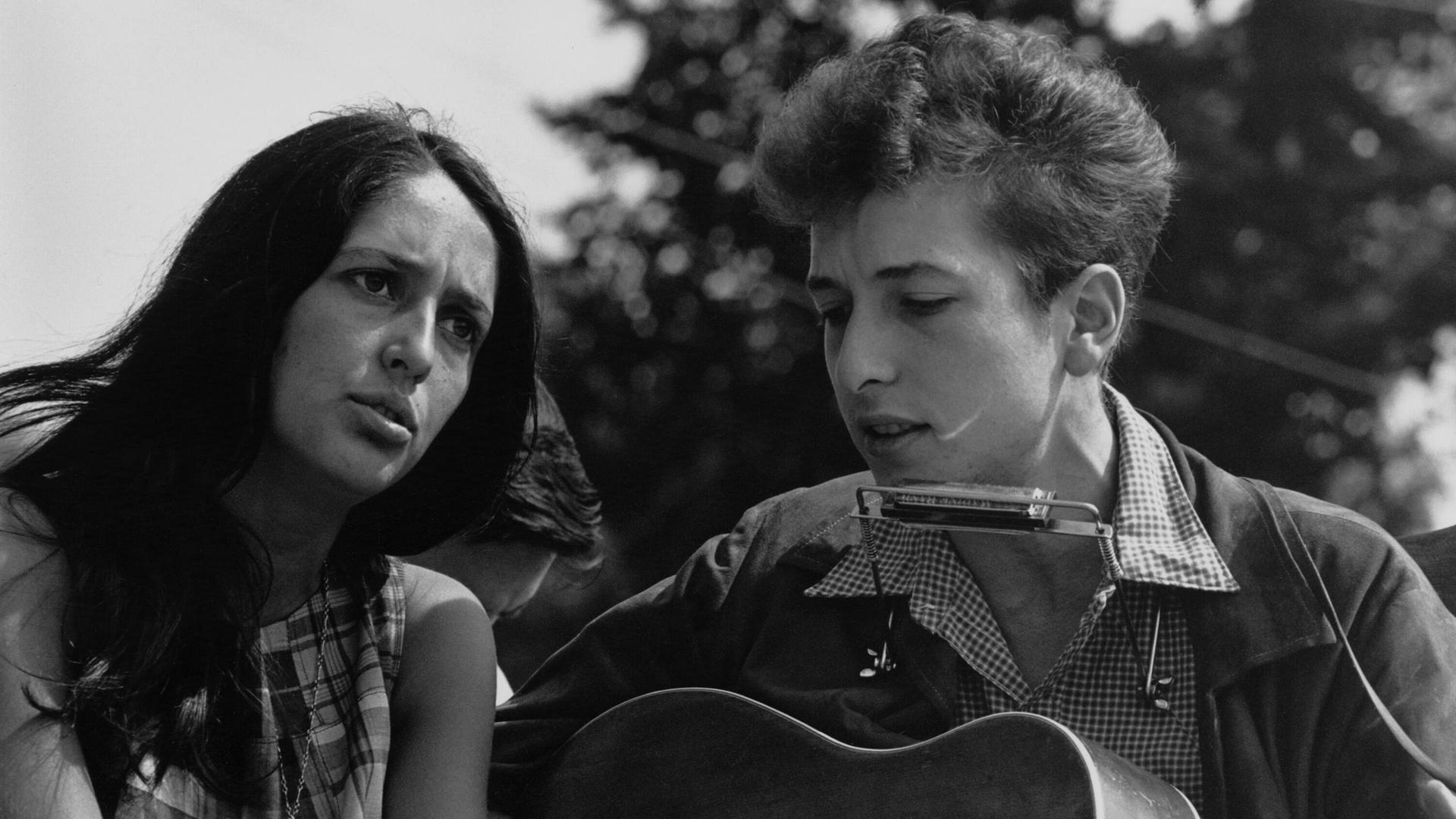 Auf dem Schwarz-Weiß-Foto sind eine dunkelhaarige weiße Frau und ein weißer Mann mit helleren Haren zu sehen. Beide singen. Der Mann spielt dabei Gitarre und hat eine Mundharmonika an einer Halterung um den Hals hängen. Es sind Joan Baez und Bob Dylan.