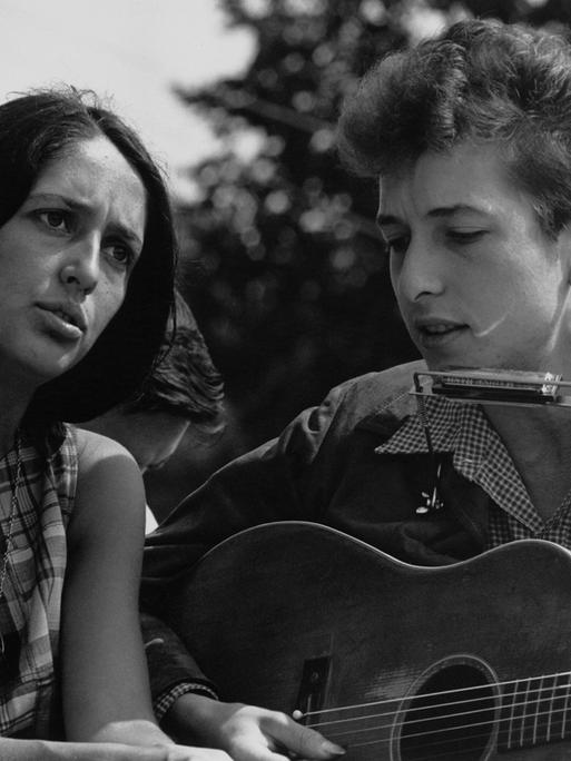 Auf dem Schwarz-Weiß-Foto sind eine dunkelhaarige weiße Frau und ein weißer Mann mit helleren Haren zu sehen. Beide singen. Der Mann spielt dabei Gitarre und hat eine Mundharmonika an einer Halterung um den Hals hängen. Es sind Joan Baez und Bob Dylan.