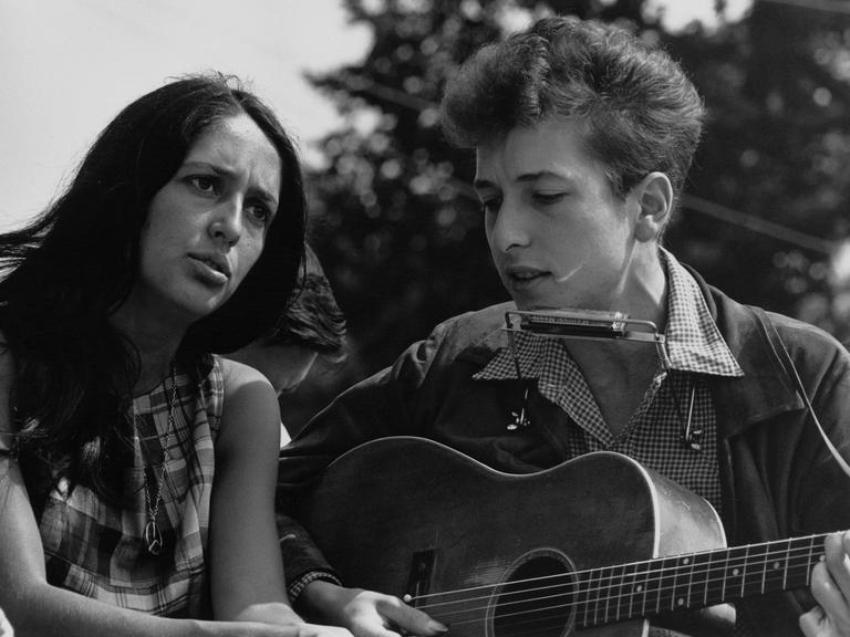 Auf dem Schwarz-Weiß-Foto sind eine dunkelhaarige weiße Frau und ein weißer Mann mit helleren Haren zu sehen. Beide singen. Der Mann spielt dabei Gitarre und hat eine Mundharmonika an einer Halterung um den Hals hängen. Es sind Joan Baez und Bob Dylan.