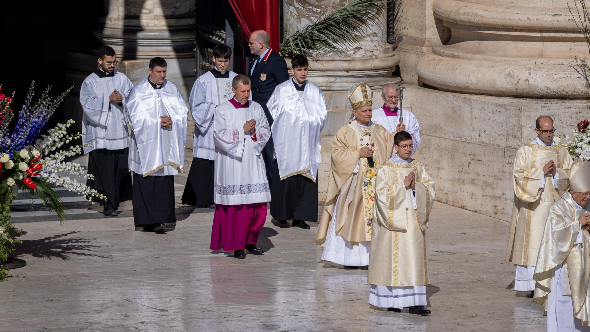 Papst Leo hat ein goldenes Gewand an. Er ist umgeben von anderen Kirchen-Männern in feierlichen Gewändern. Sie gehen über einen spiegelnden Marmor-Boden. Papst Leo hat ein goldenes Gewand an. Er ist umgeben von anderen Kirchen-Männern in feierlichen Gewändern. Sie gehen über einen spiegelnden Marmor-Boden.