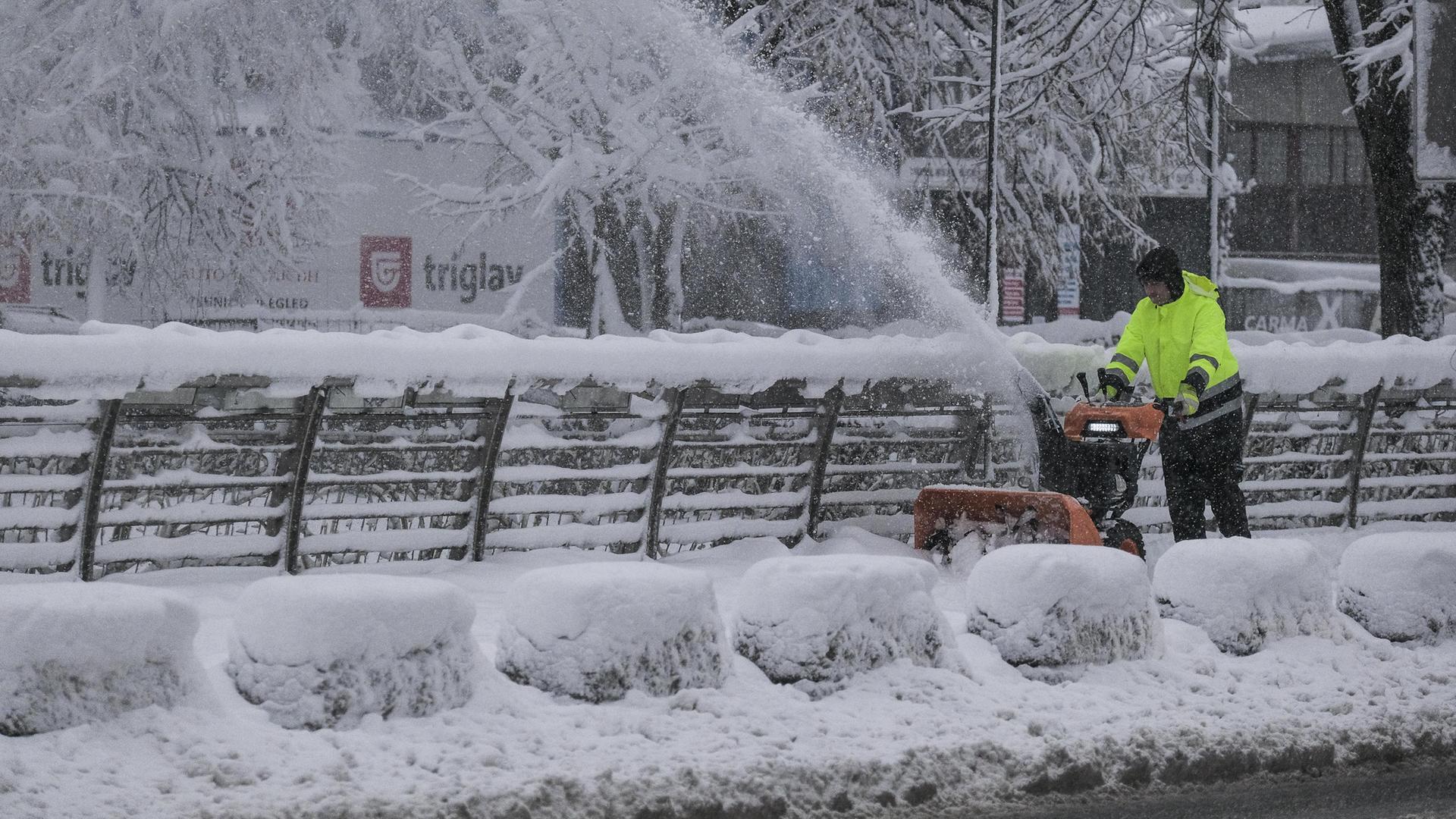 Ein Mann fährt mit einer Schneefräse über den Gehweg. Der Schnee fliegt meterhoch aus dem Gerät.