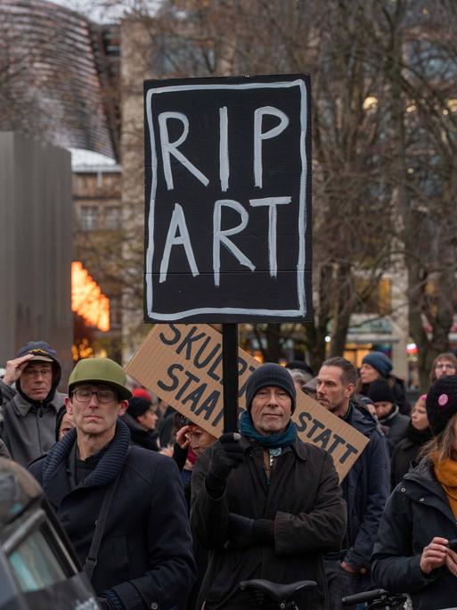 Auf einer Demo hält ein Mann ein Schild mit der Aufschrift "RIP ART" in die Luft.
