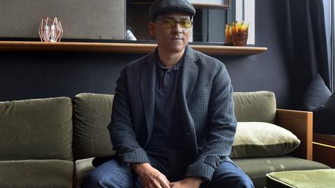 Der Sänger Xavier Naidoo sitzt auf einem Sofa und blickt in die Kamera. Er trägt eine Mütze und eine Brille mit getönten Gläsern.