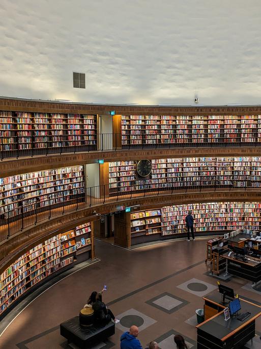 Die Stockholmer Stadtbibliothek, ein berühmtes architektonisches Wahrzeichen, verfügt über eine geräumige kreisförmige Halle mit mehreren Ebenen von Bücherregalen. Besucher stöbern in der Bibliothek, einige sitzen, andere gehen auf den Balkonen entlang. (Quelle: picture alliance)