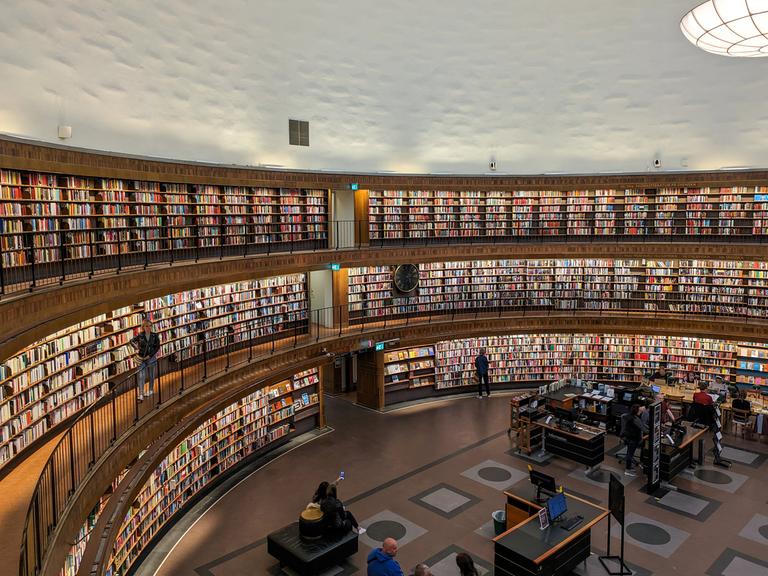 Die Stockholmer Stadtbibliothek, ein berühmtes architektonisches Wahrzeichen, verfügt über eine geräumige kreisförmige Halle mit mehreren Ebenen von Bücherregalen. Besucher stöbern in der Bibliothek, einige sitzen, andere gehen auf den Balkonen entlang. (Quelle: picture alliance)