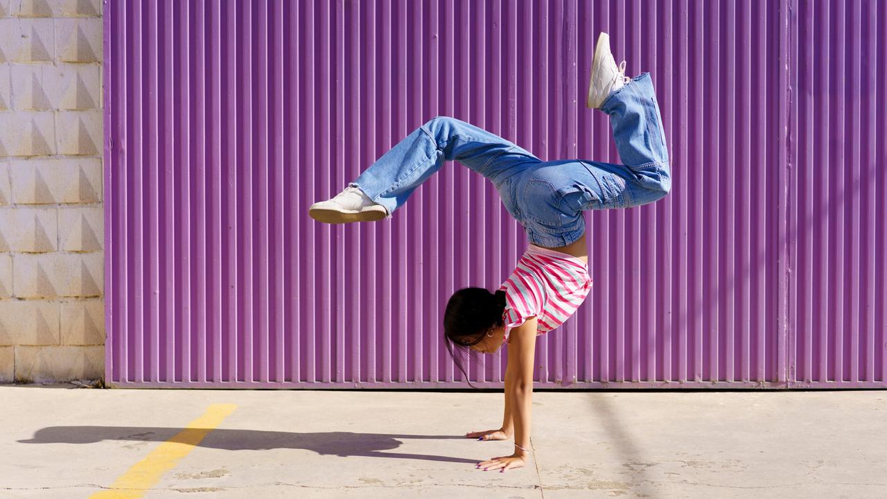 Ein Teil von Breakdance: Eine junge Frau macht vor einer violetten Wand einen Handstand. Sie trägt eine weite Jeans und Turnschuhe sowie ein gestreiftes T-Shirt.