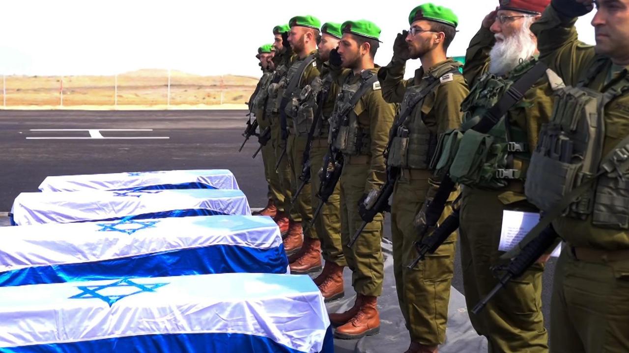 Die mit israelischen Flaggen verhüllten Särge stehen auf dem Flugfeld eines Hubschrauberlandeplatzes. Davor stehen mehrere israelische Soldaten, die salutieren. 