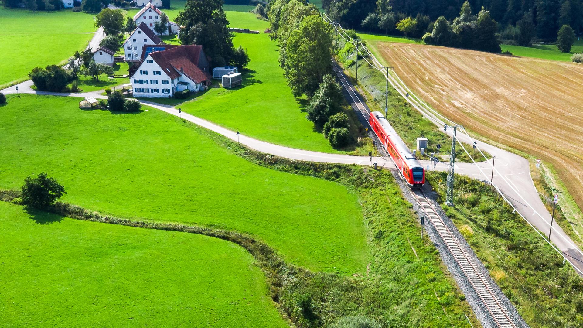 Ein roter Regionalzug fährt durch grüne Wiesen und Felder.