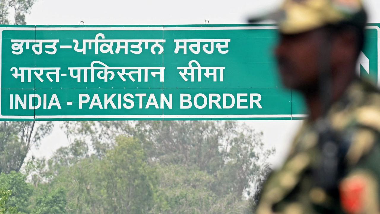 Ein grünes Schild mit weißen Buchstaben der Indisch-Pakistanischen Grenze, im Vordergrund unscharf ein Grenzposten in Uniform