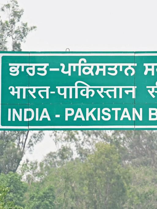 Ein grünes Schild mit weißen Buchstaben der Indisch-Pakistanischen Grenze, im Vordergrund unscharf ein Grenzposten in Uniform