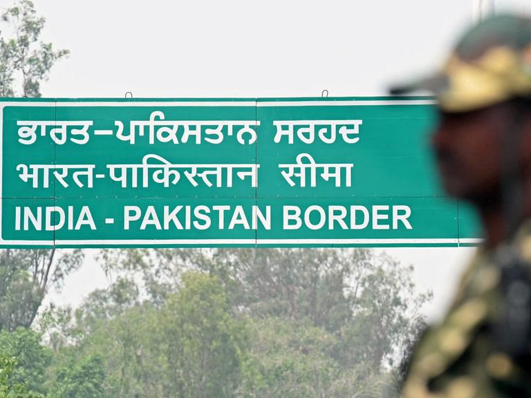 Ein grünes Schild mit weißen Buchstaben der Indisch-Pakistanischen Grenze, im Vordergrund unscharf ein Grenzposten in Uniform