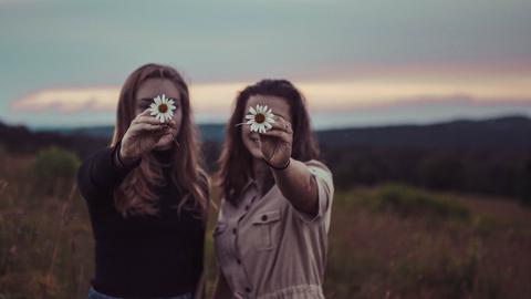 Zwei Frauen stehen vor einer wild romantischen Landschaft mit untergehender Sonne und halten lachend jeweils eine weiße Blume so vor das Gesicht, dass nur die Blumen scharf erscheinen.