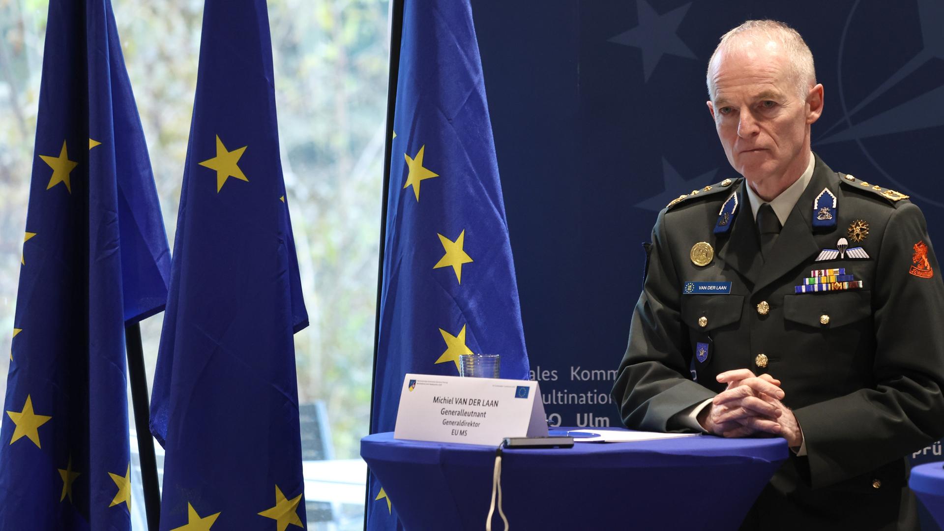 Van der Laan steht ernst an einem Rednerpult. Seine Hände sind ineinander verschränkt, hinter ihm sind drei EU-Flaggen. 