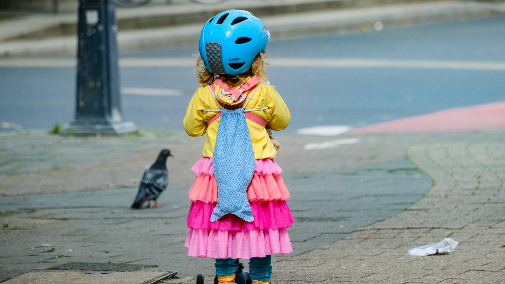 Ein kleines Kind mit einem Tretroller, einer gelben Jacke und einem pinken Rock. Auf dem Kopf hat das Kind einen blauen Helm. Auf dem Rücken einen Rucksack in Form eines blauen Fisches.