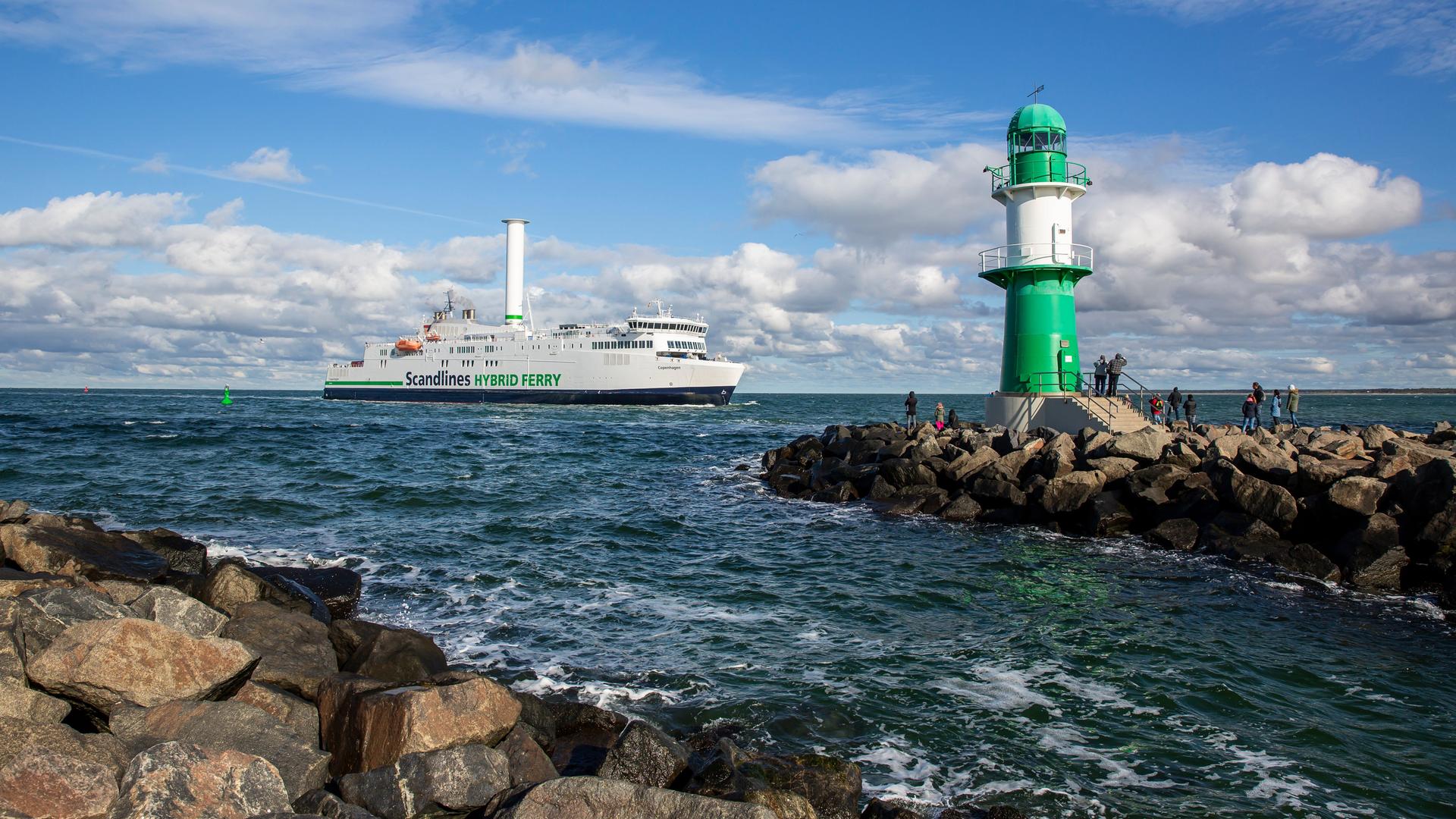 Die Scandlines Hybrid Fähre Copenhagen mit ihrem Rotorsegel bei der Einfahrt in den Hafen von Warnemünde. Im Vordergrund das grün-weisse Westmolenfeuer.