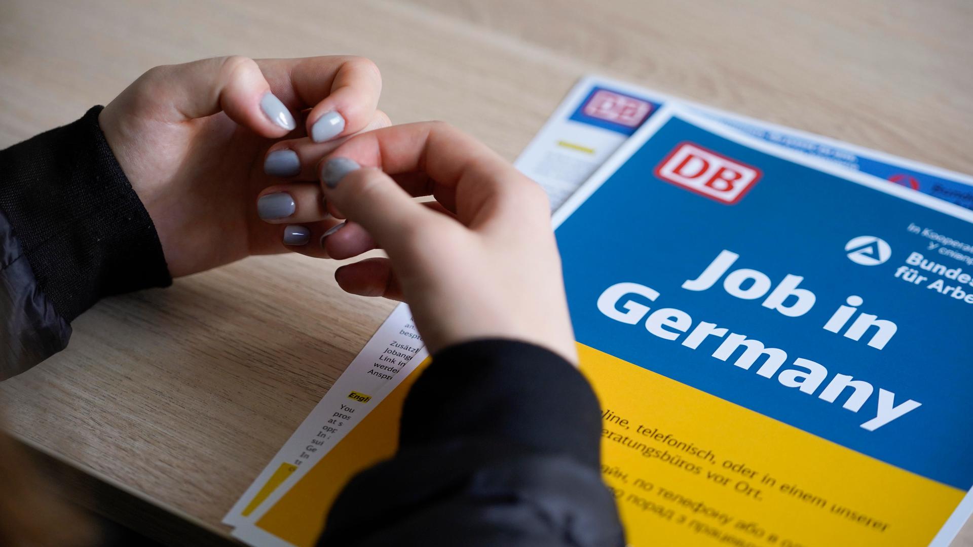 Die Hände einer Frau mit taubenblauem Nagellack liegen auf einem Informationsblatt der Deutschen Bahn, es ist in den ukrainischen Nationalfarben Blau und Gelb gehalten und trägt die Aufschrift "Job in Germany".