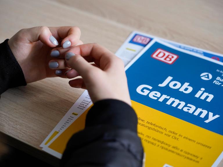 Die Hände einer Frau mit taubenblauem Nagellack liegen auf einem Informationsblatt der Deutschen Bahn, es ist in den ukrainischen Nationalfarben Blau und Gelb gehalten und trägt die Aufschrift "Job in Germany".