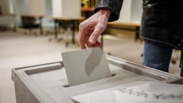 Das Foto zeigt eine Hand beim Einwerfen eines Stimm-Zettels in eine Wahl-Urne.